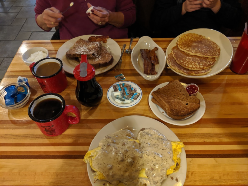 A table set with breakfast items: pancakes, bacon, eggs with gravy, toast, syrup, and coffee mugs.