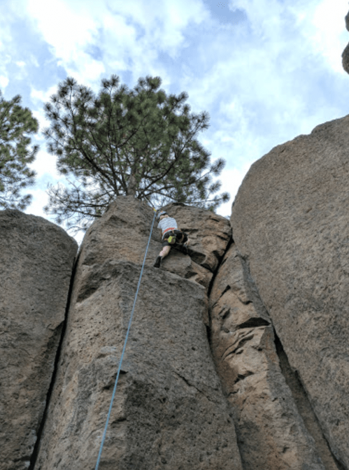A climber ascends a rocky cliff, with a tree visible above and a blue sky in the background.