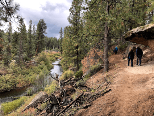 Two people walk along a dirt path beside a river, surrounded by trees and rocky terrain under a cloudy sky.