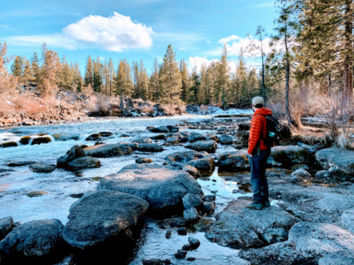A person in a red jacket stands on rocky riverbanks, surrounded by trees and a clear blue sky.