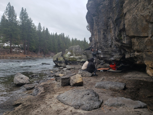 A group of people gathered near a river, sitting on rocks and under a cliff, surrounded by trees and a cloudy sky.