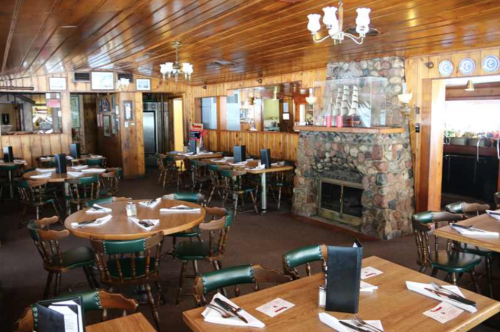 Cozy restaurant interior with wooden walls, stone fireplace, round tables, and green chairs, set for dining.