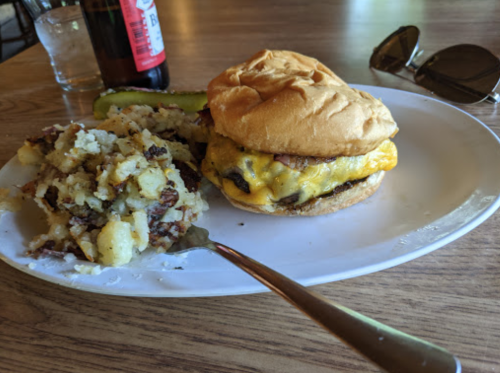 A plate with a cheeseburger, a side of potato salad, a pickle, and a drink in the background. Sunglasses nearby.