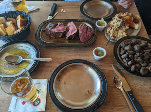 A table set with steak, mashed potatoes, mushrooms, bread, and a beer, with empty plates and utensils.