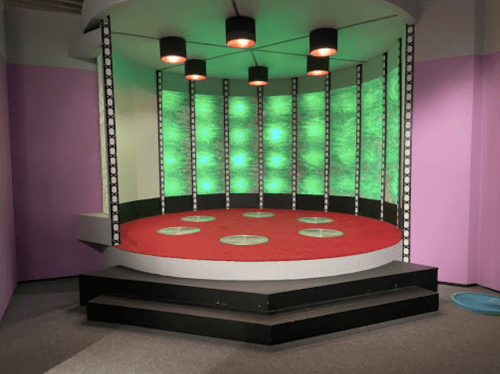 A futuristic stage with a circular red platform, green lights, and black accents, set against a pink wall.