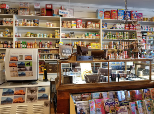 A cozy store interior filled with shelves of canned goods, snacks, and magazines, featuring a display case with leather items.