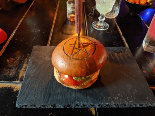 A burger with a pentagram design burned into the bun, served on a slate plate in a dimly lit setting.