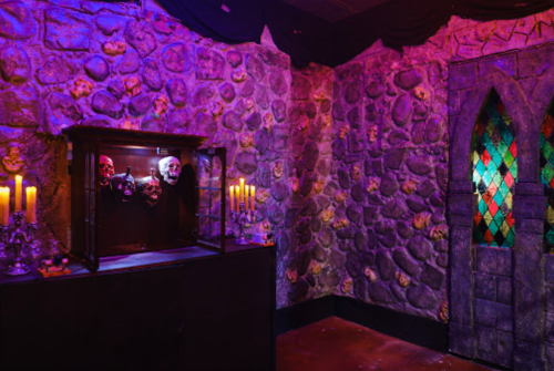 A dimly lit room with stone walls, featuring a display of skulls, candles, and a stained glass window.