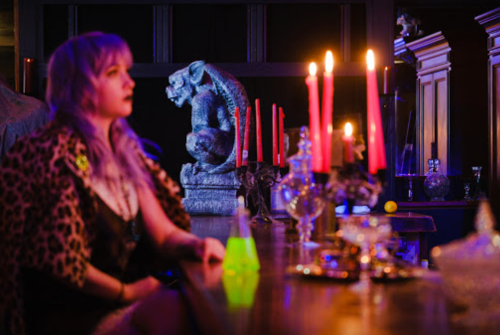 A woman with purple hair sits at a dimly lit bar, with candles and a gargoyle statue in the background.