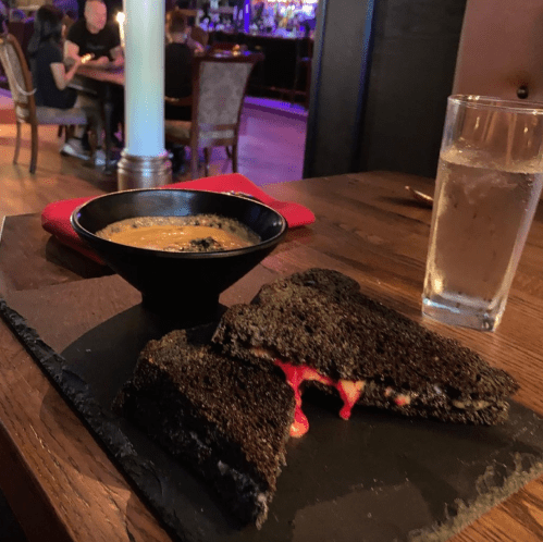 A black sandwich with melted cheese, served on a slate plate alongside a bowl of soup and a glass of water.