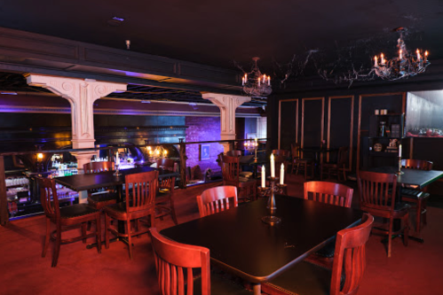 A dimly lit bar with wooden tables, chairs, and chandeliers, featuring a stylish, intimate atmosphere.