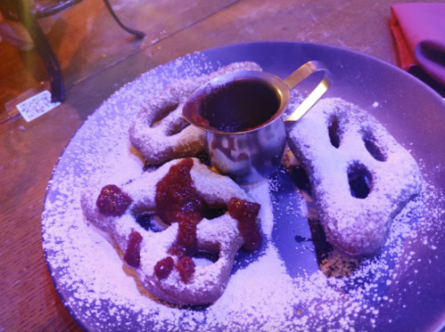 A plate of powdered sugar-dusted pastries shaped like pretzels, served with a small jug of chocolate sauce.