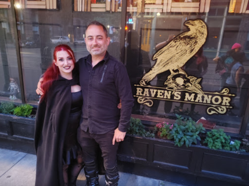 A woman in a black outfit and a man in a black shirt pose together outside Raven's Manor, with a raven logo in the background.