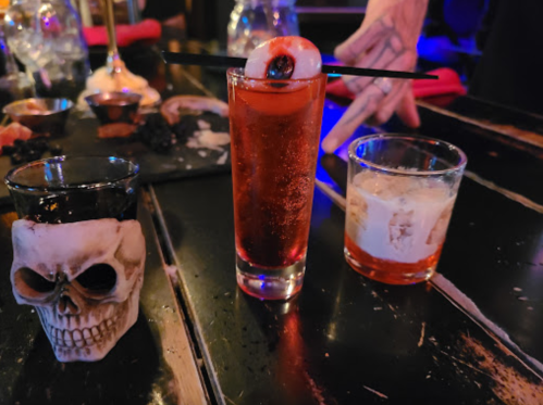 Three cocktails on a bar: a red drink with an eyeball garnish, a black drink in a skull glass, and a whiskey on the rocks.