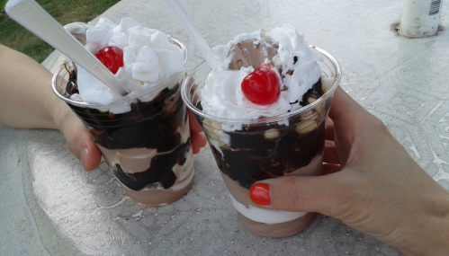 Two hands holding ice cream cups topped with whipped cream and cherries, set on a table outdoors.