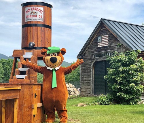A bear mascot in a green hat stands with arms outstretched near a wooden mining structure and a rustic building.
