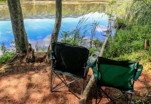 Two camping chairs sit by a calm lake, surrounded by trees and greenery, reflecting the blue sky above.