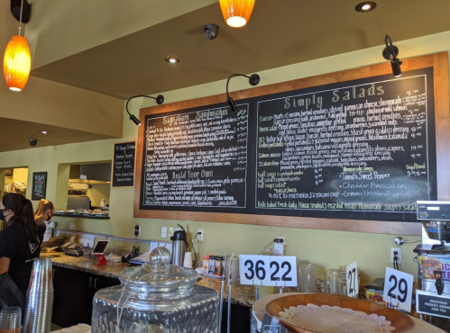 A café menu board featuring salad options and daily specials, with staff members in the background.