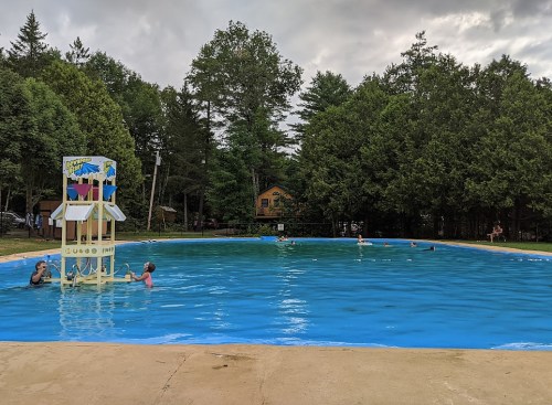 A swimming pool surrounded by trees, with people swimming and playing near a water play structure.
