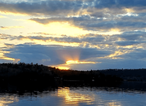 A serene sunset over a calm lake, with clouds reflecting warm hues and a silhouette of trees in the foreground.