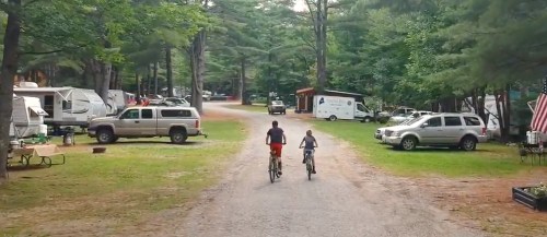 Two children ride bicycles down a gravel path in a wooded campground, surrounded by parked vehicles and trees.