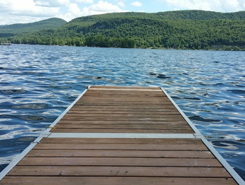 A wooden dock extends into a calm lake, surrounded by green hills and a blue sky with fluffy clouds.