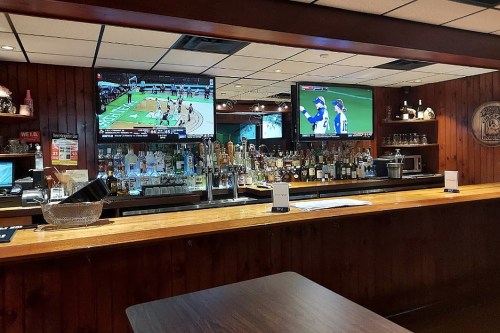 A bar with a wooden counter, shelves of liquor, and two TVs showing sports games in a cozy setting.