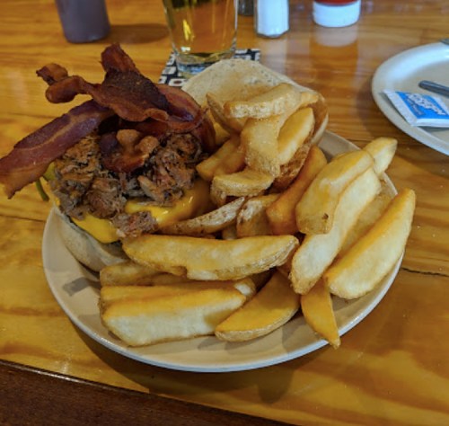A plate with a loaded burger topped with bacon, cheese, and pulled pork, served with crispy fries.