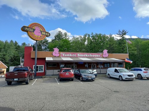 Exterior of Yankee Smokehouse with a sign featuring a pig, surrounded by parked cars and trees in the background.