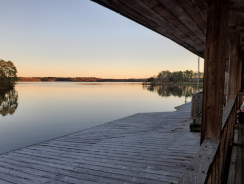 A serene lake view at sunrise, with a wooden dock extending into calm waters and trees lining the shore.