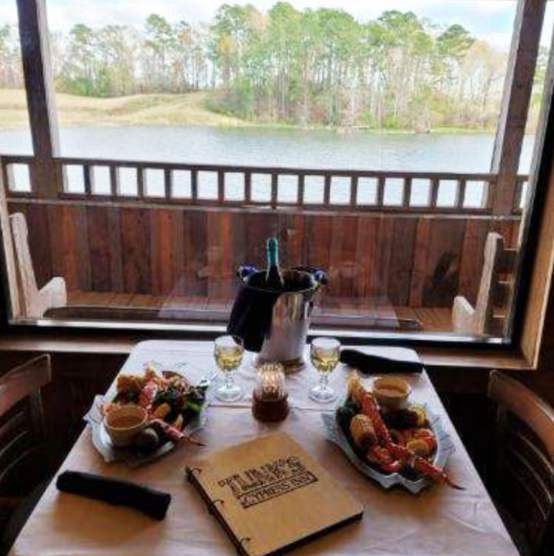A dining table with seafood dishes and drinks, overlooking a serene lake through a wooden window.