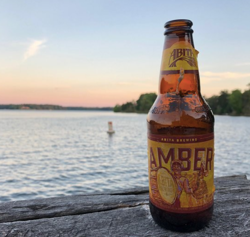 A bottle of Abita Amber beer sits on a wooden surface by a lake at sunset, with trees in the background.