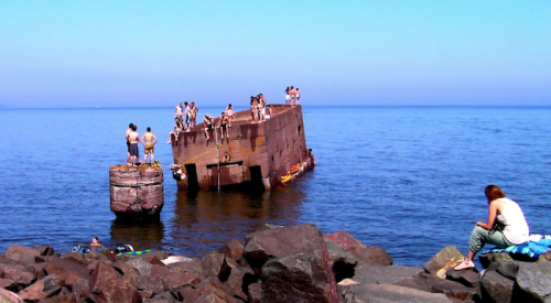 People enjoying a sunny day by the water, some on a concrete structure, others relaxing on rocks.