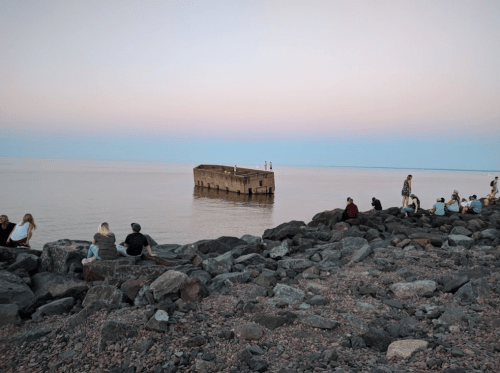 People sit on rocky shorelines at sunset, with a sunken structure partially visible in calm waters.
