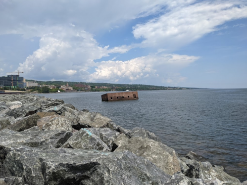 A partially submerged, rusted structure in calm water, with rocky shoreline and clouds in the background.