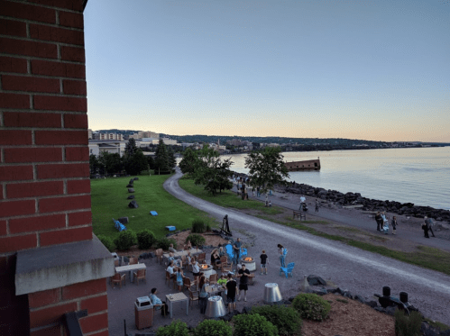 A scenic view of a waterfront area with people gathered around a fire pit, grassy spaces, and a calm shoreline at dusk.