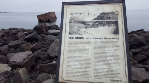 A sign about "The Cribs" near a sunken structure by the water, with rocky shoreline and calm lake in the background.