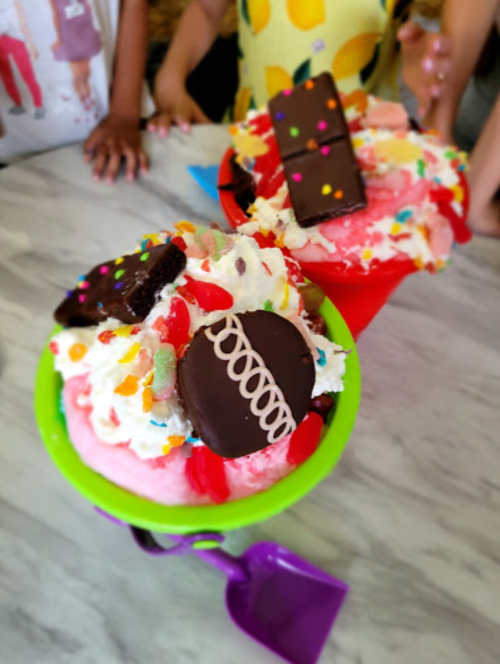 Two colorful ice cream bowls topped with whipped cream, chocolate, sprinkles, and candy, with children's hands in the background.