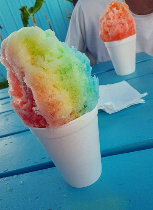 Two colorful snow cones in white cups, one with rainbow hues and the other in shades of red and orange, on a blue table.