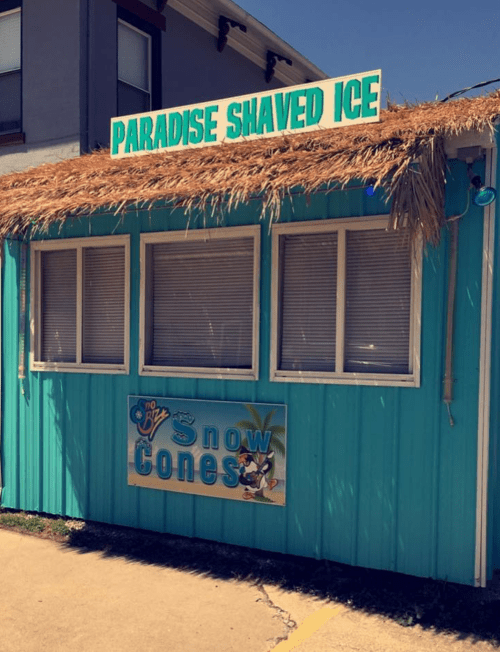 A colorful shack with a thatched roof, labeled "Paradise Shaved Ice," offering snow cones.