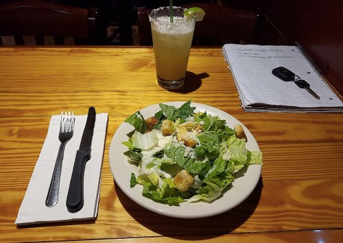 A fresh salad with lettuce, croutons, and a drink on a wooden table, with a notebook and keys in the background.