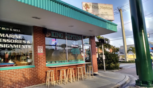 Exterior of King Neptune restaurant with a green awning, bar stools outside, and a sign above. Palm trees in the background.