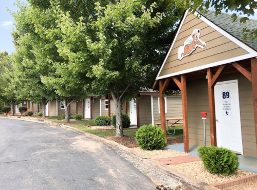 A row of small cabins with green trees and shrubs lining a paved path, featuring a dog logo on one cabin.