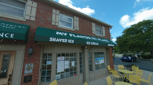Exterior of a brick building with green awnings reading "My Flavor It Is Place" and outdoor seating.