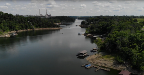 A serene river scene with boats along the shore, surrounded by greenery and distant industrial structures.