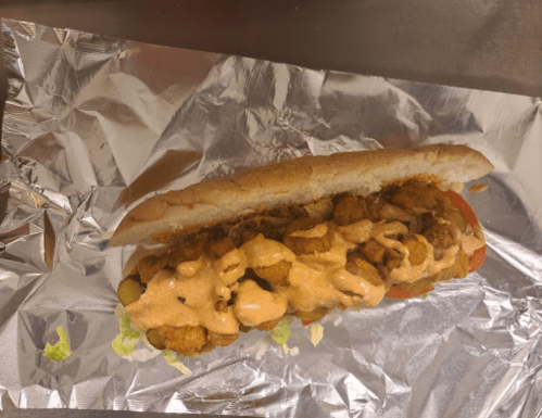 A sub sandwich filled with fried shrimp, lettuce, tomato, and drizzled with a spicy sauce, wrapped in foil.