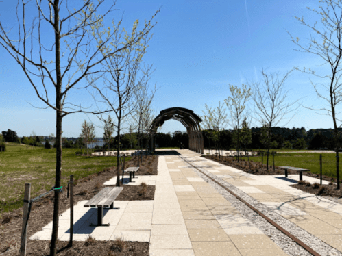A sunny park pathway lined with trees and benches, leading to a wooden arch structure.