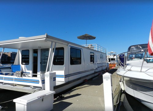 A white houseboat docked at a marina, with a blue sky and other boats nearby.