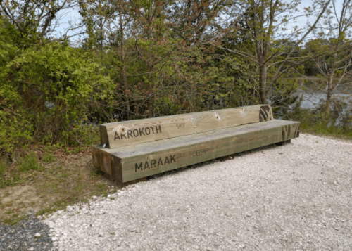 A wooden bench with the names "Akrokoth" and "Maraak" carved into it, surrounded by greenery near a water body.