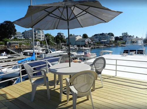 A patio with a table and chairs under an umbrella, overlooking a marina filled with boats and waterfront homes.